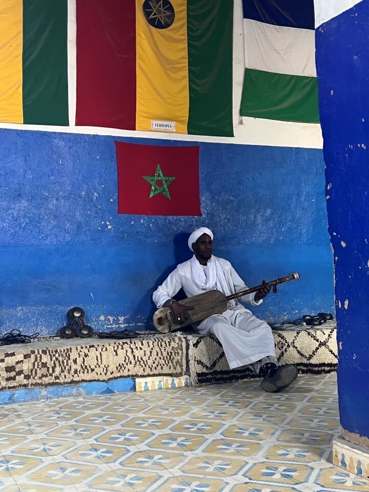 hombre negro con instrumento musica con túnica blanca sentado en un banco con paredes azules y madera de marruecos colgada y otras banderas africanas en marruecos