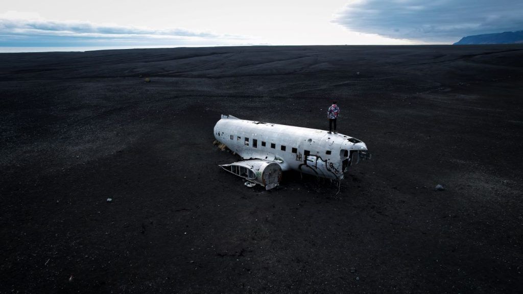 vistas desde arriba avion abandonado sin alas en playa negra infinita y chico subido encima del avion mirando de espaldas al horizonte