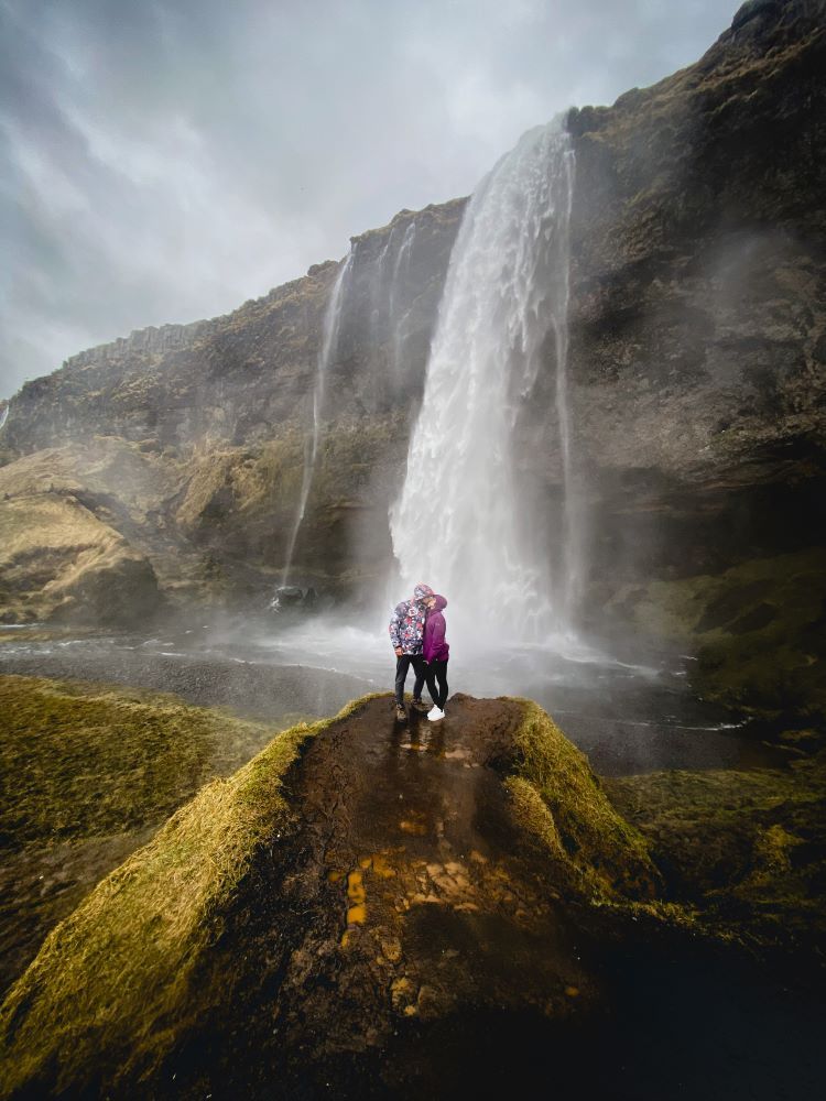 cascada de fondo y dos personas con chaquetas de color lila y parches blanco besandose en la boca y posando de frente delante de la cascada en Islandia