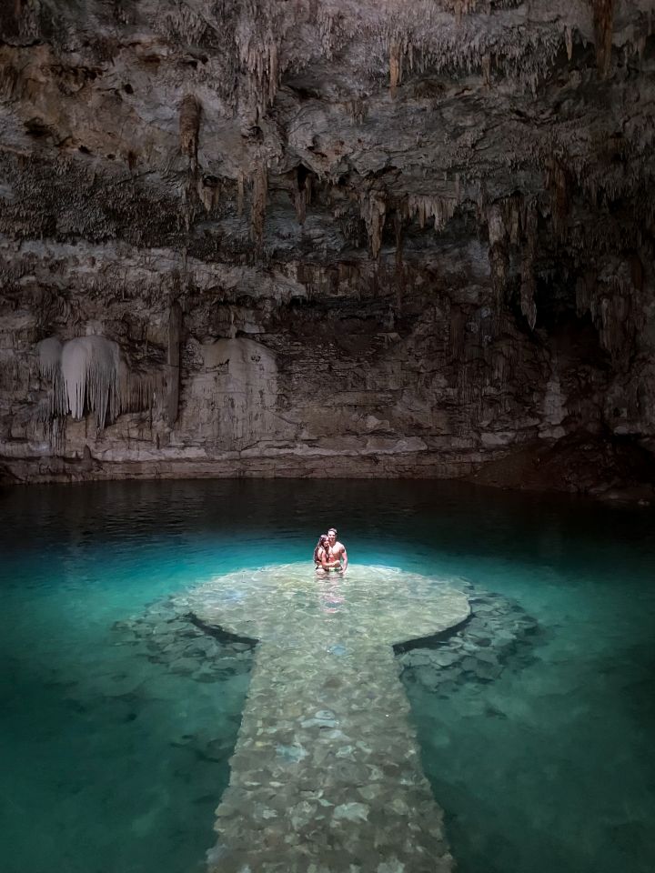 cueva con pasarela dentro del agua y donde personas abranzandose al final donde les ilumina un rayo de sol que sale del techo