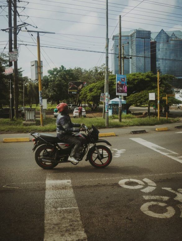 carretera con moto parado, con edificios de fondo y motorista es negro con gorro de lana naranja en la cabeza
