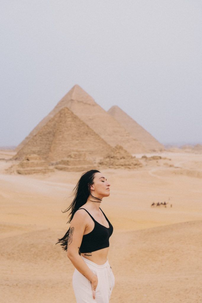 piramides de egipto de fondo y chica de pelo largo moreno con top negro de tirantes y pantalon blanco de lino mirando hacia la derecha con los ojos cerrados y viento en la cara