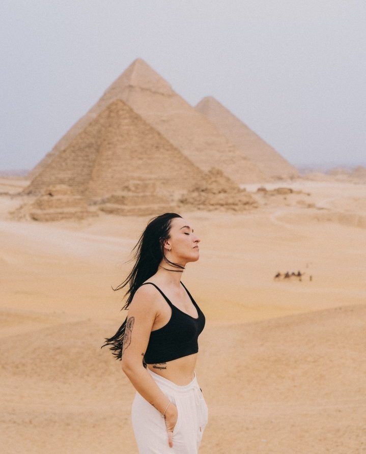 piramides de egipto de fondo y chica de pelo largo moreno con top negro de tirantes y pantalon blanco de lino mirando hacia la derecha con los ojos cerrados y viento en la cara