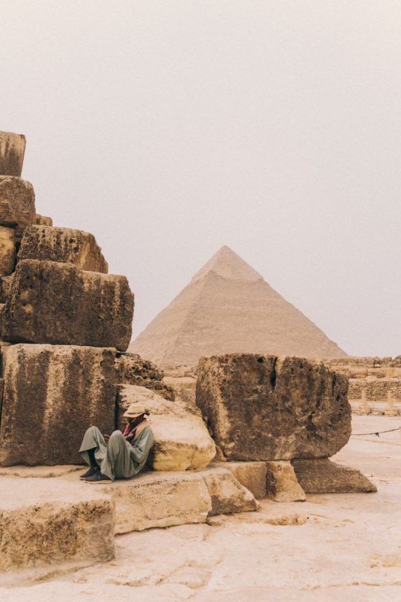 hombre tumbado y apoyado en una piedra junto algunas piedras mas y la pirámide de Egipto de fondo