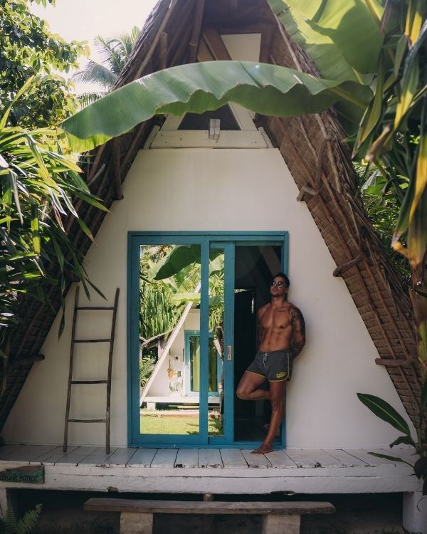 chico apoyado en la puerta de un cabaña con bañador con gafas de sol mirando a la cámara
