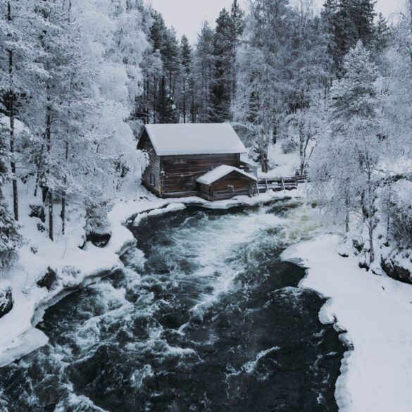 paisaje nevado con rio que cruza y casa de madera nevada de fondo