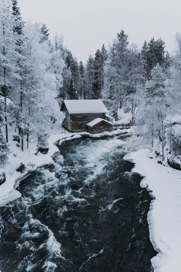 paisaje nevado con rio que cruza y casa de madera nevada de fondo